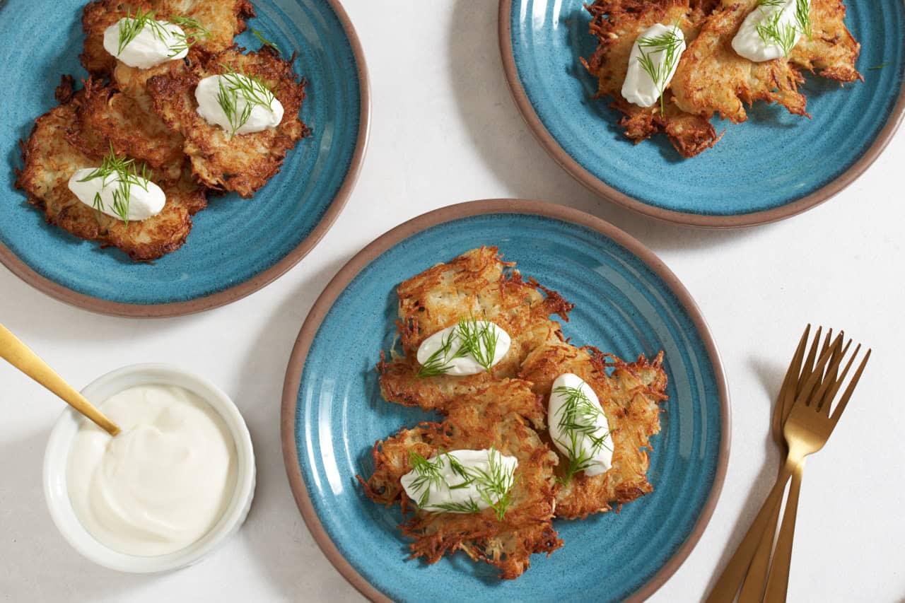 Ukrainian potato pancakes (deruny) topped with sour cream and dill on 3 blue plates, gold forks are on the right, a bowl of sour cream is on the left.