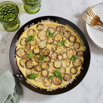 A zucchini frittata in a cast iron pan, two green glasses and a stack of white plates with gold forks on it are above it.