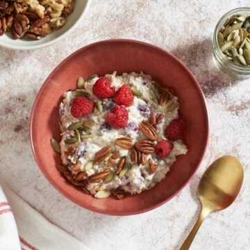 A bowl of bircher muesli topped with raspberries and nuts, surrounded by a gold spoon, small bowls of nuts and seeds, a gold spoon and a pink glass.