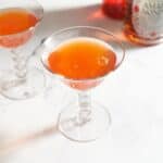 Two paper plane cocktails with bottles of liquor in the background.
