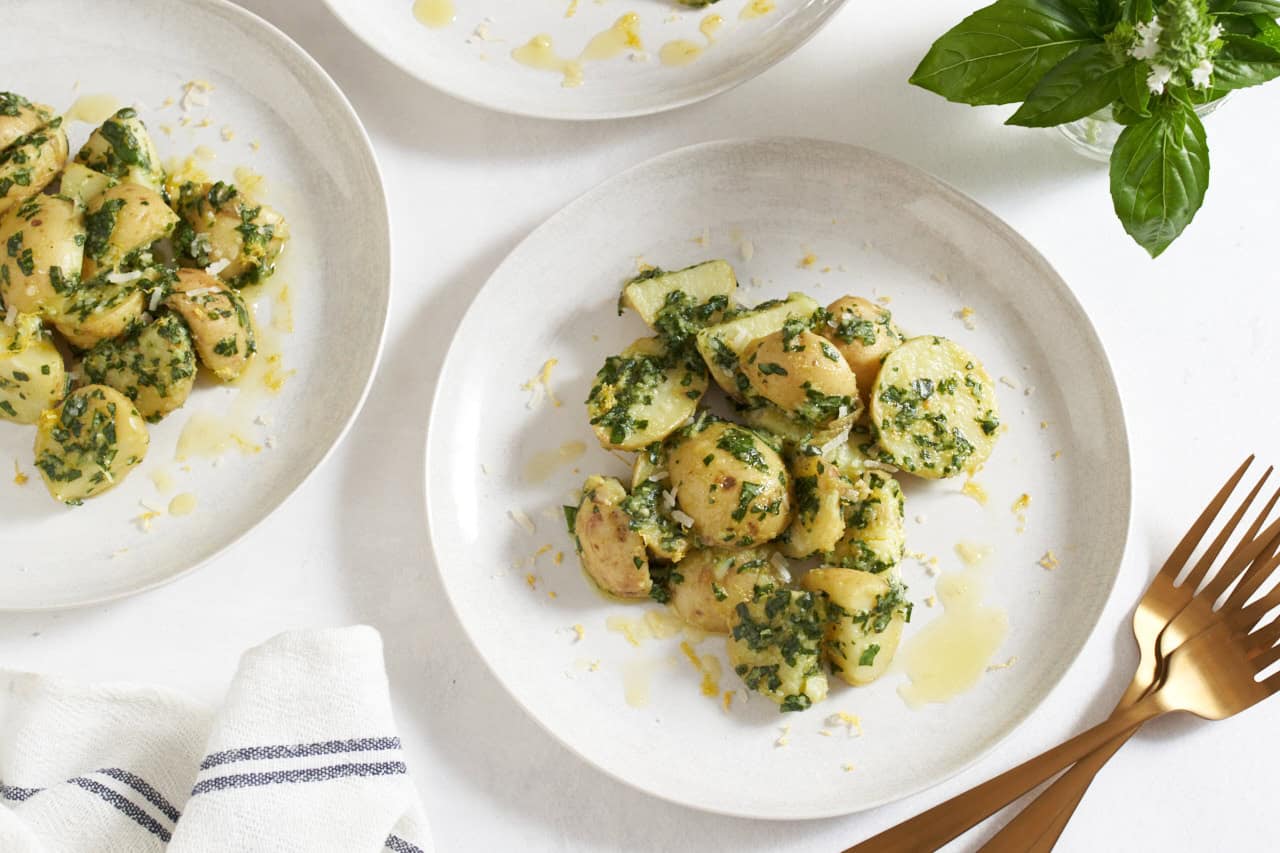 Two white plates with pesto potato salad, a jar of fresh basil is in the upper right, three gold forks are in the bottom right.