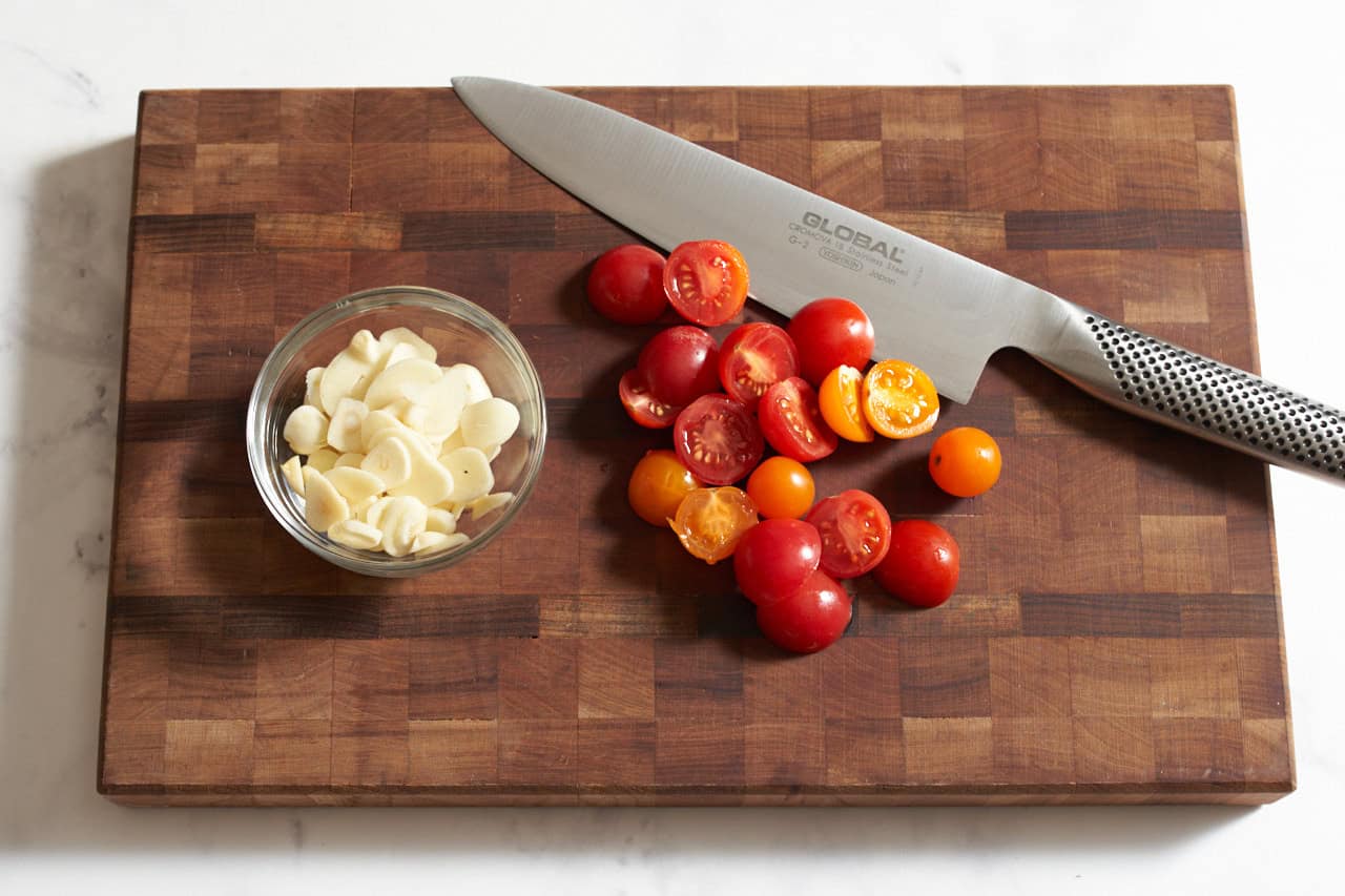 A knife on a cutting board with sliced cherry tomatoes and a bowl of sliced garlic.