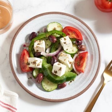 A plate of Greek salad, a glass of rosé wine, some tomatoes, a gold fork, and a striped towel surround the plate.