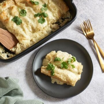 Chicken enchiladas suizas on a black plate topped with fresh cilantro, a casserole dish of enchiladas is in the top left, two gold forks are on the right.