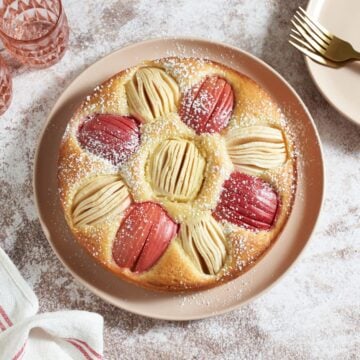 German apple cake topped with powdered sugar on a pink plate, two pink glasses are in the upper left, pink plates and gold forks are on the right, a red and white striped towel is on the bottom left.