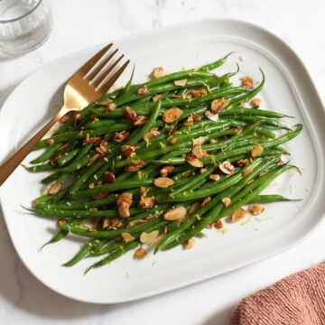 A platter of green beans amandine with a gold serving fork on it, an orange towel is on the bottom right, two glasses of water are in the upper left.