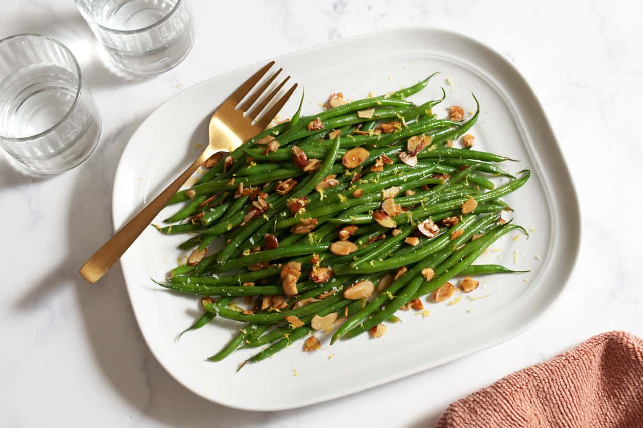 A platter of green beans amandine with a gold serving fork on it, an orange towel is on the bottom right, two glasses of water are in the upper left.