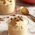 Two small servings of pumpkin mousse in glass jars, gold spoons and a large red bowl of mousse ar in the upper right.