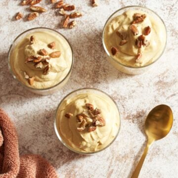 Three glasses filled with pumpkin mousse and topped with chopped pecans, a gold spoon is in the bottom right, more pecans are in the upper left.