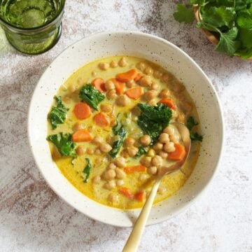 A bowl of chickpea stew with a gold spoon in it, a green glass and a bowl of cilantro are above it.