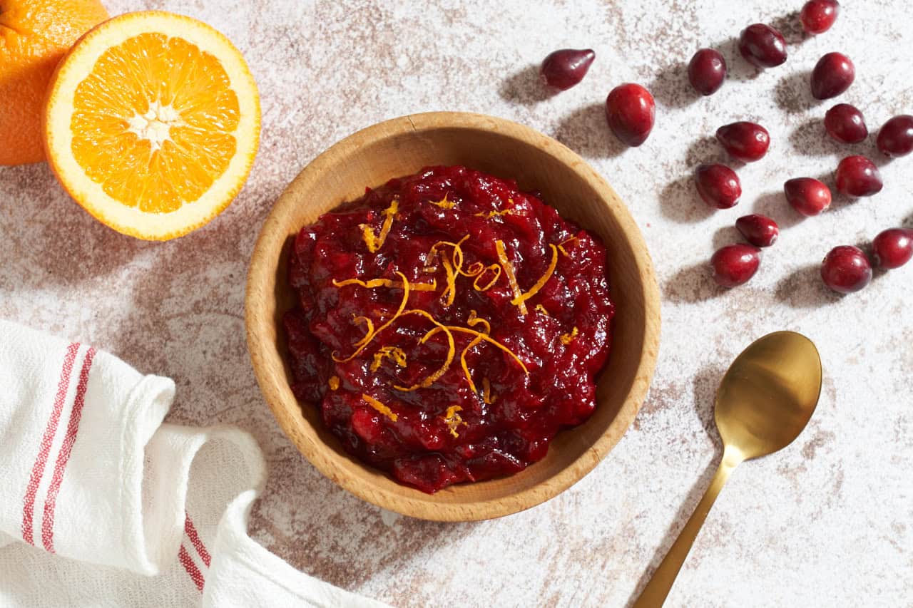 A bowl of cranberry compote topped with orange zest, half an orange, fresh cranberries, a gold spoon and a red and white striped towel surround the bowl.