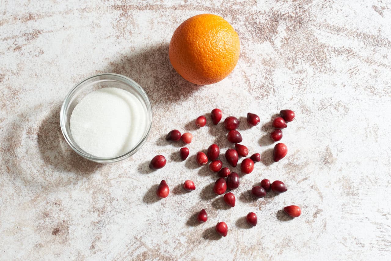 Fresh cranberries, an orange, and a bowl of sugar.