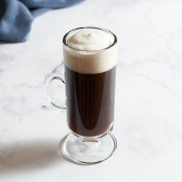 A mug of Irish coffee topped with whipped cream on a marble surface, a blue towel is in the background.