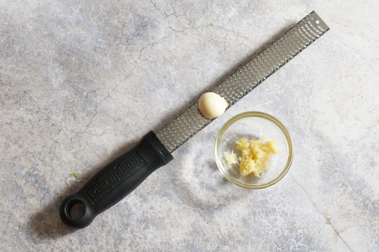 A garlic clove on a microplane grater with a small bowl of grated garlic next to it.