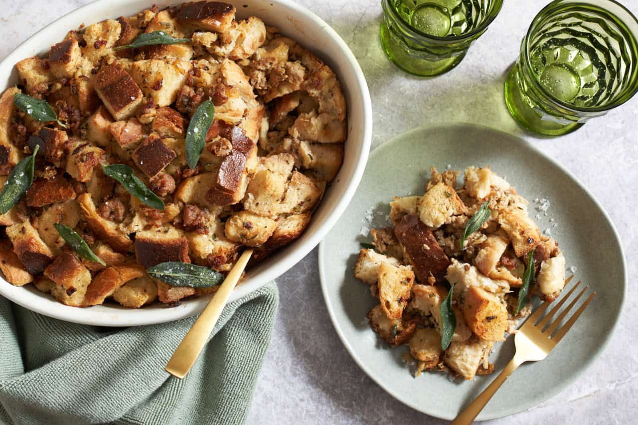 Italian sausage stuffing topped with fried sage leaves in a white casserole dish, a plate of stuffing with a gold fork is on the right, two green glasses are in the upper right.