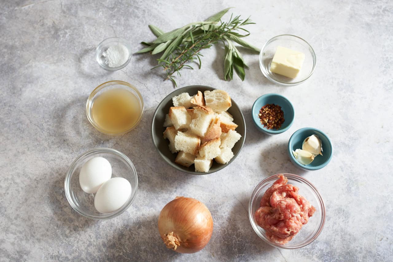 An onion, fresh sage and rosemary, and small bowls of eggs, italian sausage, cubes of bread, garlic cloves, crushed red pepper, butter, salt, and chicken stock.