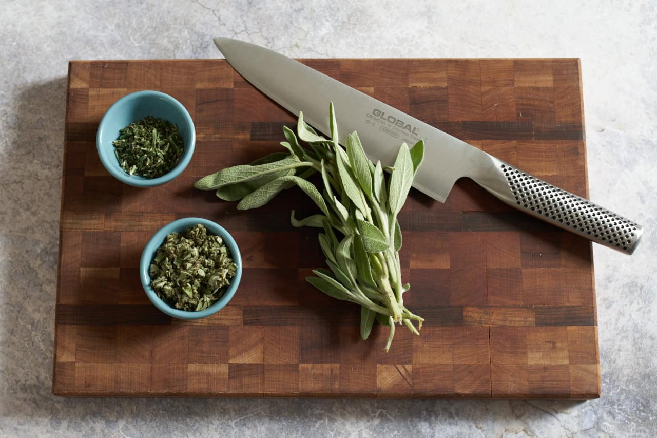 A knife on a cutting board with fresh sage and small blue bowls of chopped sage and rosemary.