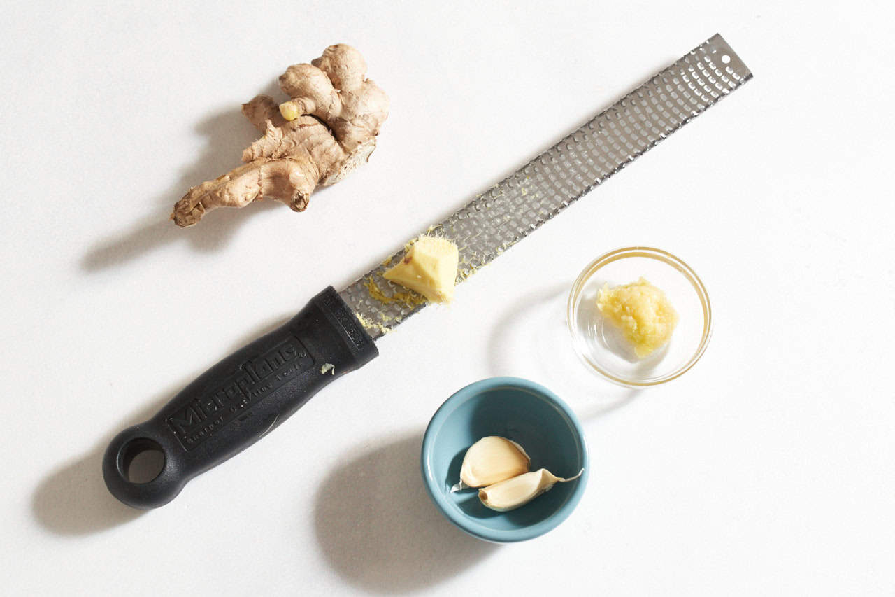 A stem of ginger, a piece of ginger on a microplane grater, and small bowls of grated garlic and garlic cloves.