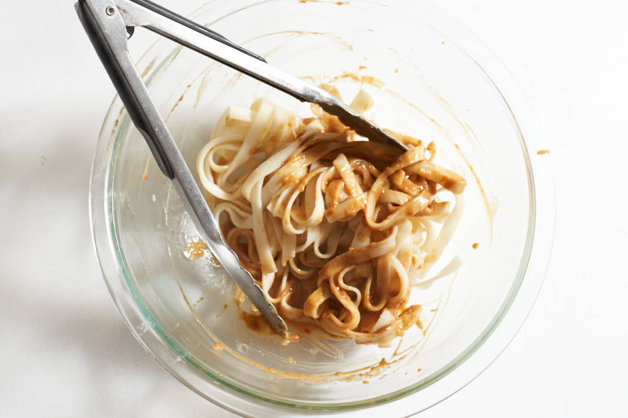 Tongs with noodles in a bowl being tossed with spicy peanut butter sauce.