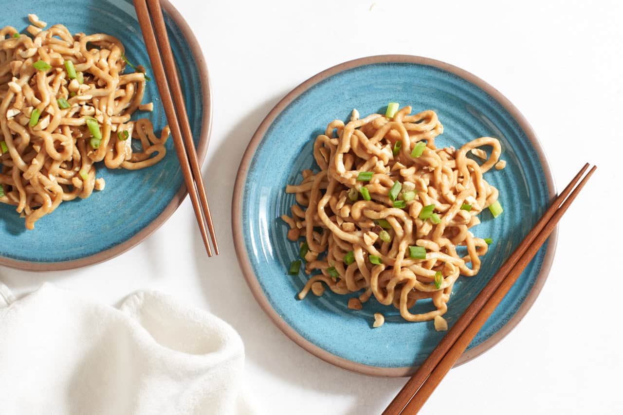 Spicy peanut noodles on two blue plates topped with chopped peanuts and scallions, with chopsticks on the side.