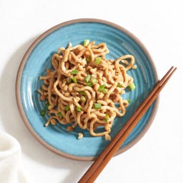 Spicy peanut noodles on a blue plate topped with chopped peanuts and scallions, with chopsticks on the side.