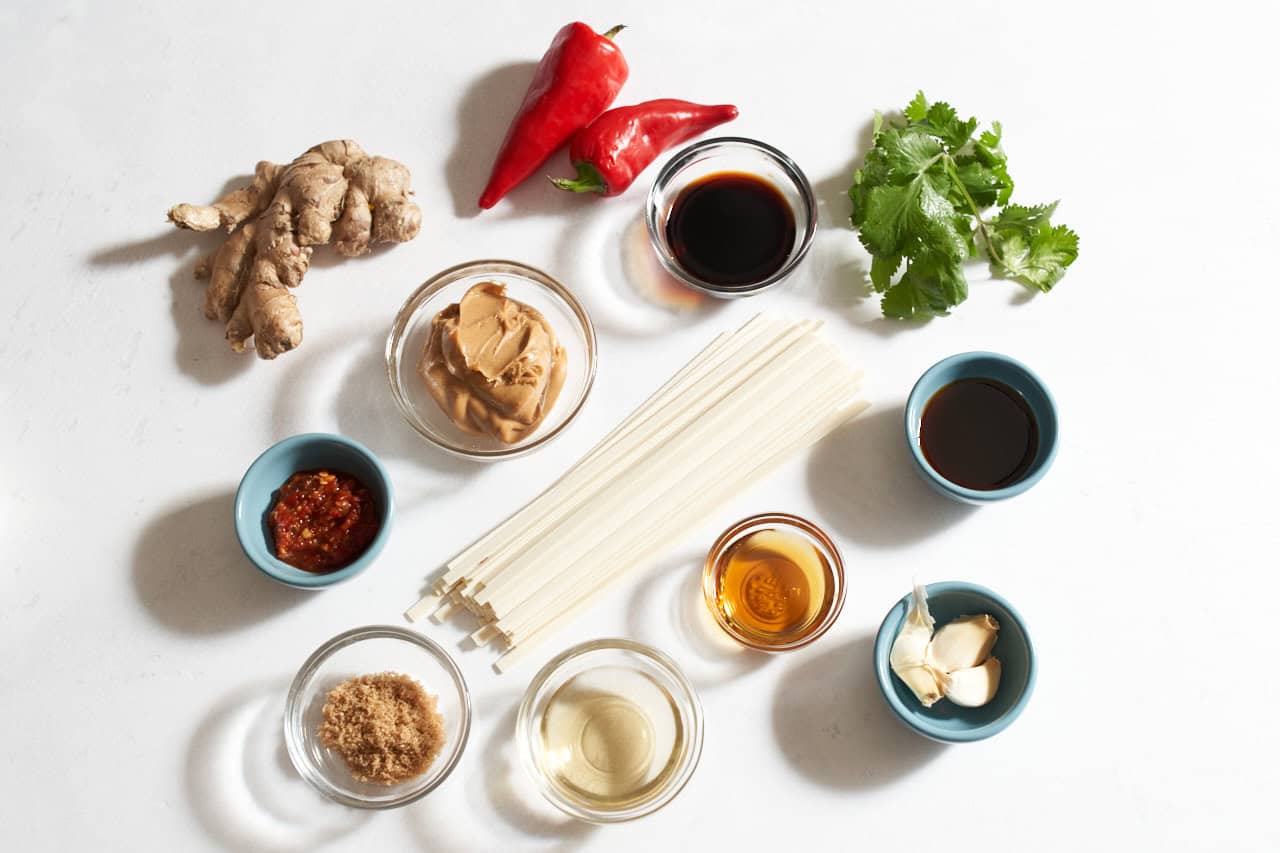 Fresh ginger, chili peppers, fresh cilantro, noodles and small bowls of peanut butter, soy sauce, sesame oil, fish sauce, brown sugar, garlic-chile sauce, and garlic cloves.