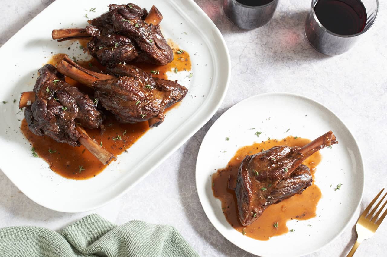 A platter of red wine braised lamb shanks and sauce next to a single lamb shank on a white plate, two glasses of red wine are in the upper right, a gold fork is on the bottom right.