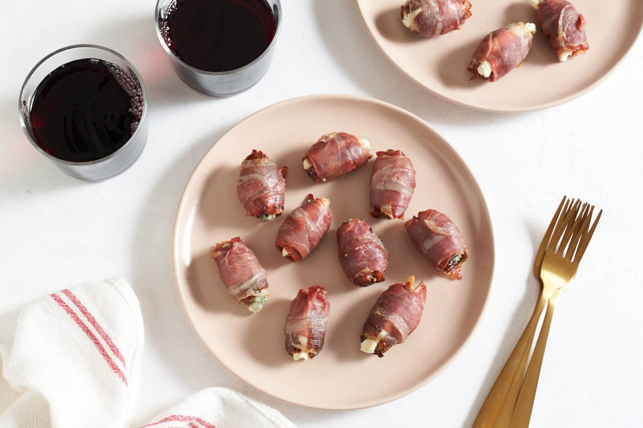 Two pink plates of prosciutto wrapped dates, gold forks are on the right, two glasses of red wine are in the upper left.