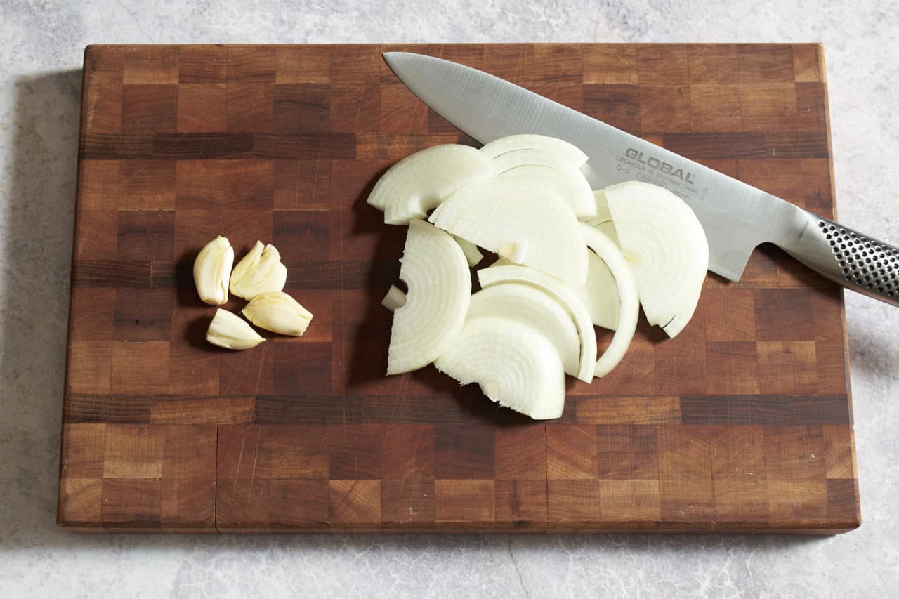 A knife on a cutting board with sliced onions and smashed garlic cloves.