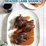 Braised lamb shanks and red wine sauce on a white platter, a plate with a single lamb shank and two glasses of red wine are above it.