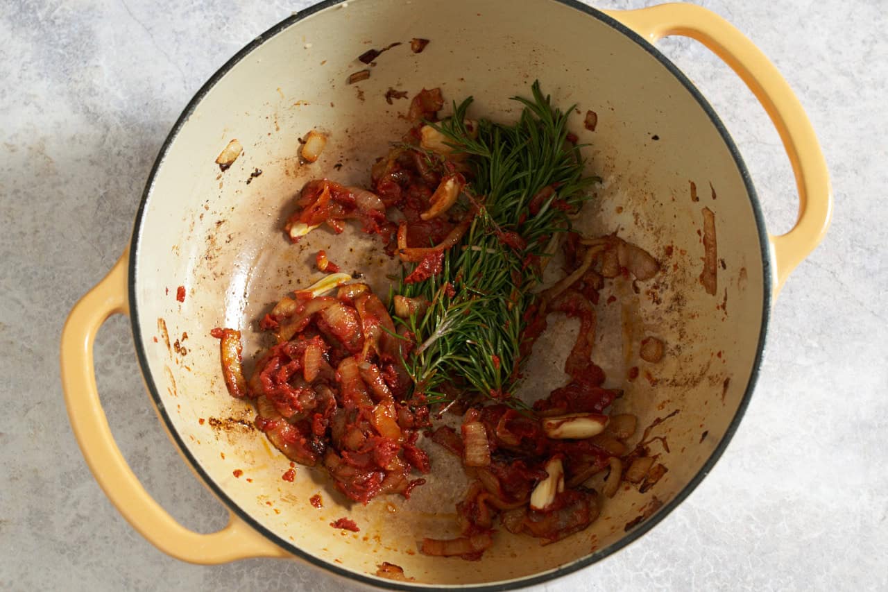 Onions, garlic, tomato paste, and a bundle of fresh rosemary and thyme in a yellow dutch oven.