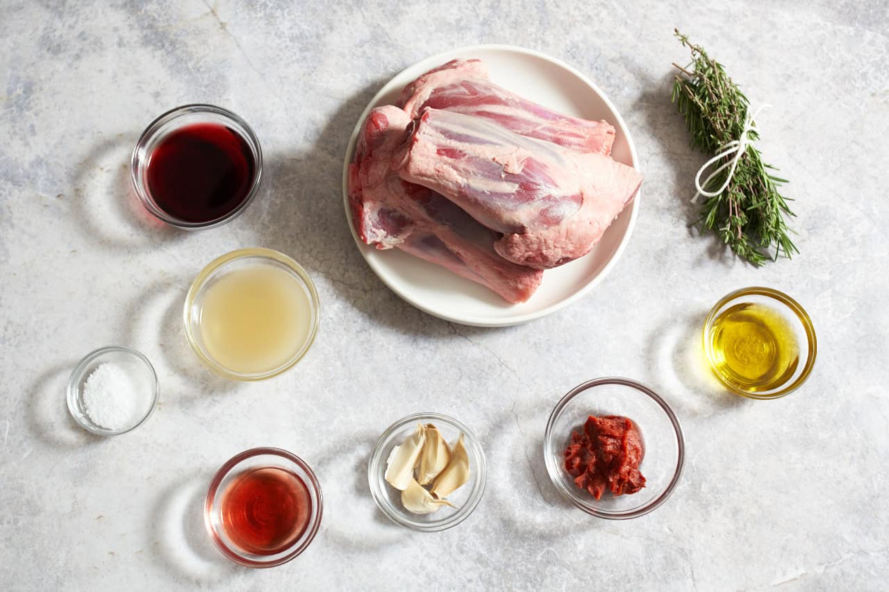 Raw lamb shanks on a plate surrounded by small bowls of olive oil, tomato paste, red wine, red wine vinegar, garlic cloves, chicken stock, kosher salt, and bundle of rosemary and thyme.