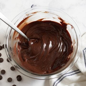 A bowl of dark chocolate ganache with a spoon in it, chocolate chips are on the left, a blue and white towel is on the right.