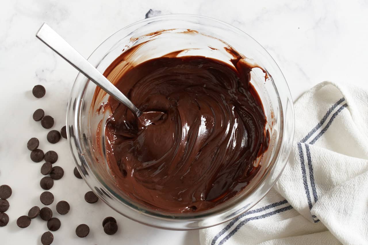 A bowl of dark chocolate ganache with a spoon in it, chocolate chips are on the left, a blue and white towel is on the right.