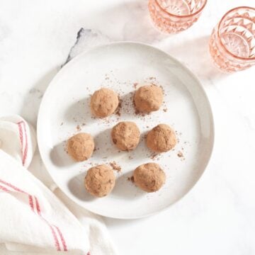 Dark chocolate ganache truffles coated in cocoa powder on a white plate, two pink glasses are in the upper right, a red and white striped towel is in the lower left.