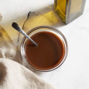 A small bowl of dijon balsamic vinaigrette with a spoon in it, a bottle of olive oil is in the upper right, a brown towel is on the bottom left.