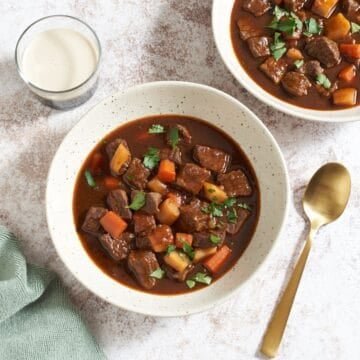 Two bowls of Guinness beef stew topped with parsley, a glass of Guinness beer is on the left, a gold spoon is on the right.