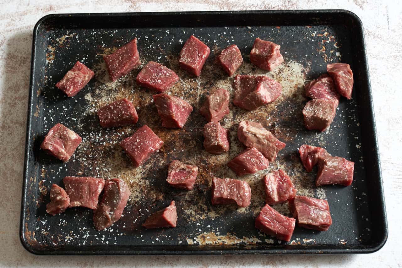 Beef stew meat cut into smaller chunks on a sheet pan seasoned with kosher salt and pepper.
