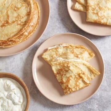 Two Ukrainian crepes with a sweet cheese filling folded on a pink plate, two more plates of stacked and folded crepes are above it, a bowl of filling is on the left.