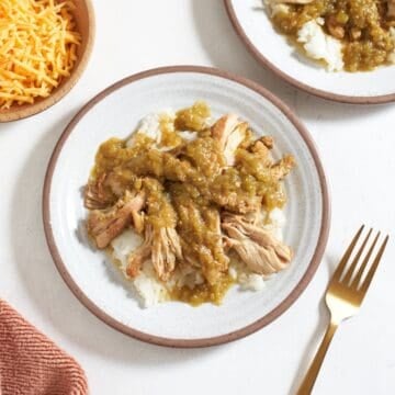 Two plates of slow cooker green chili chicken served over rice, a wooden bowl of shredded cheese is on the left, a gold fork is to the right.