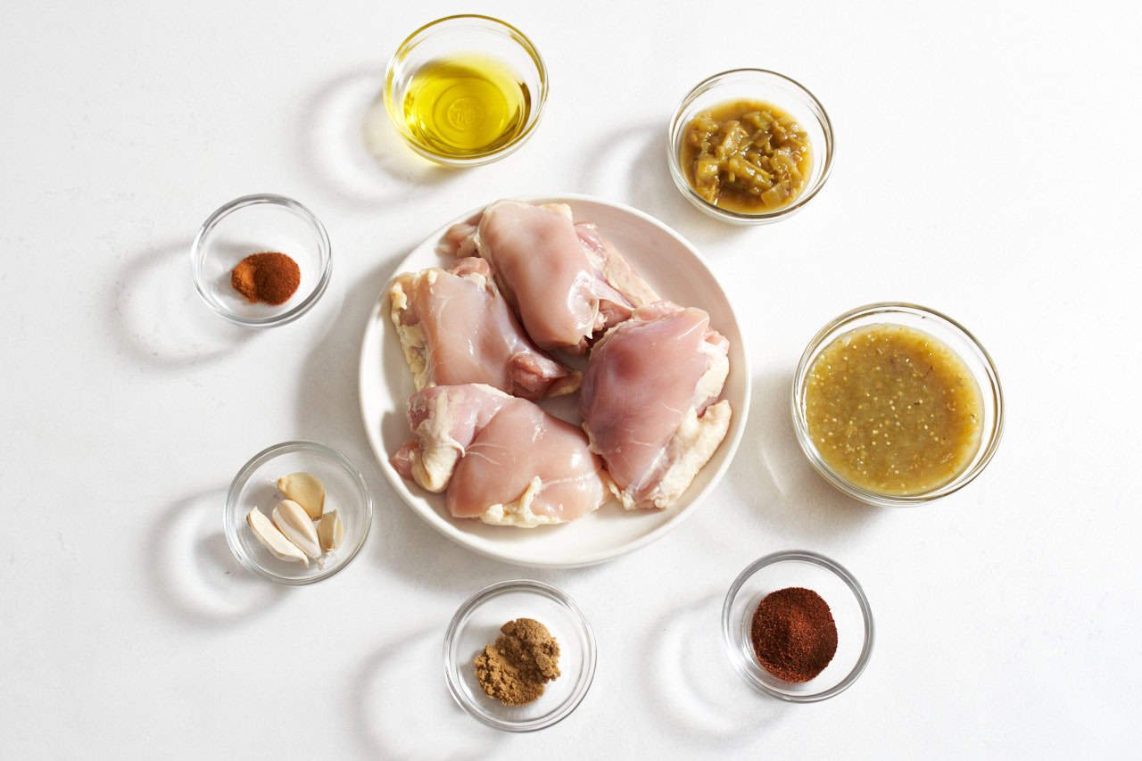 Chicken thighs on a white plate surrounded by small bowls of chili powder, green chilis, salsa verde, garlic, olive oil, and spices.