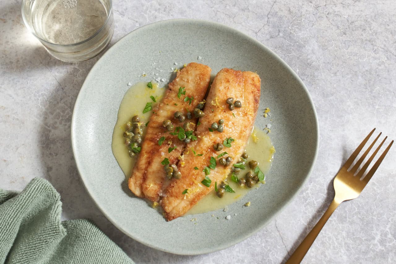 A plate of fish with lemon butter sauce on top, garnished with fresh parsley, a gold fork is on the right, a glass of white wine is in the upper left.