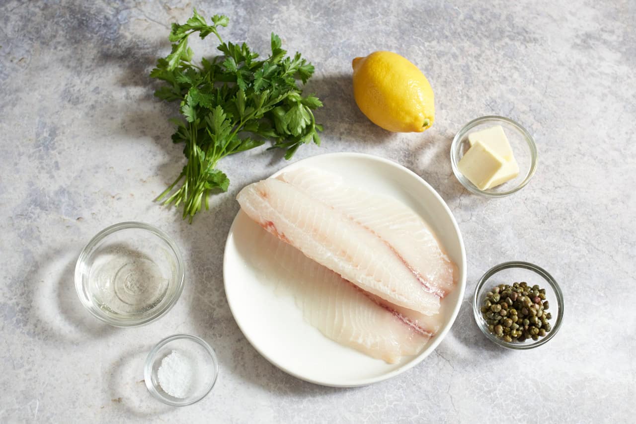 Fresh parsley, a lemon, a plate of fish fillets, and small bowls of capers, butter, white wine, and salt.