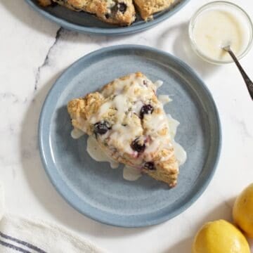 A glazed lemon blueberry scone on a blue plate, a small bowl of lemon glaze is in the upper right corner.