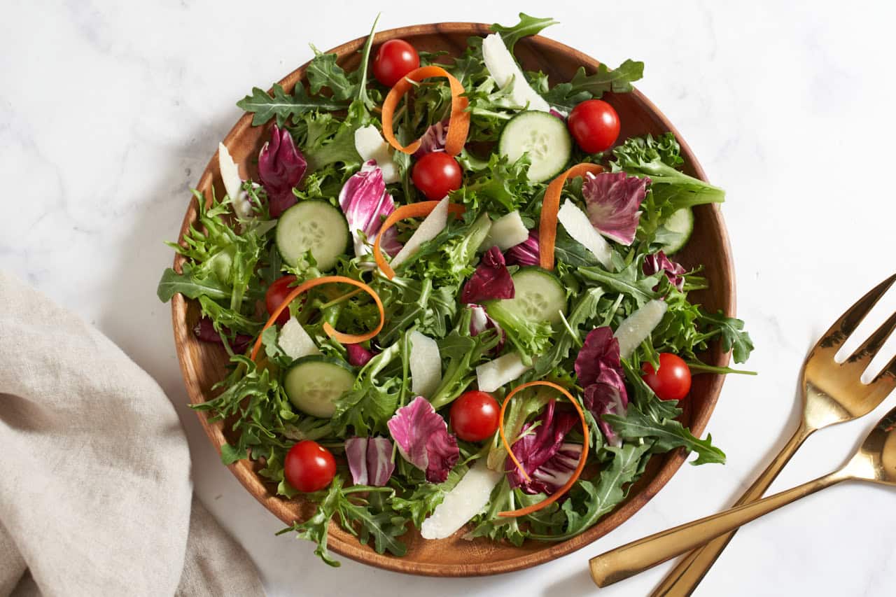 An insalata mista (Italian mixed salad) on a wooden plate, gold serving utensils are on the right.