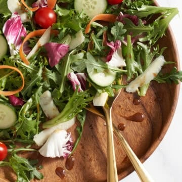 An insalata mista (Italian mixed salad) on a wooden plate with gold serving utensils.