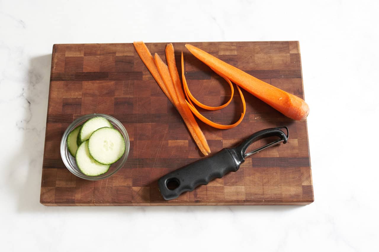 Grated carrots, a small bowl of sliced cucumbers, and a vegetable peeler on a wooden cutting board.