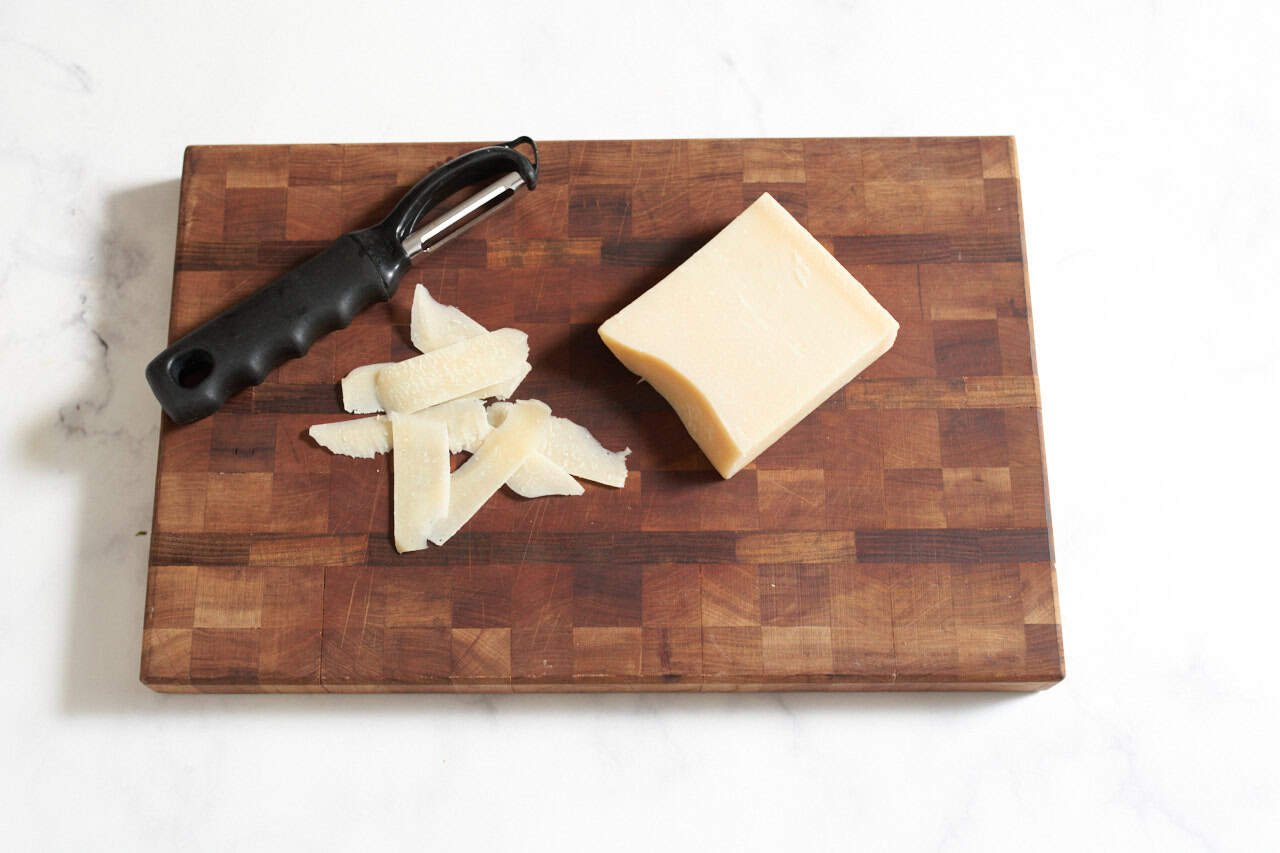 A block of parmesan, some parmesan shavings and a vegetable peeler on a wooden cutting board.