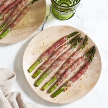 Two plates of baked prosciutto wrapped asparagus topped with lemon zest, a green glasses is in the upper middle.