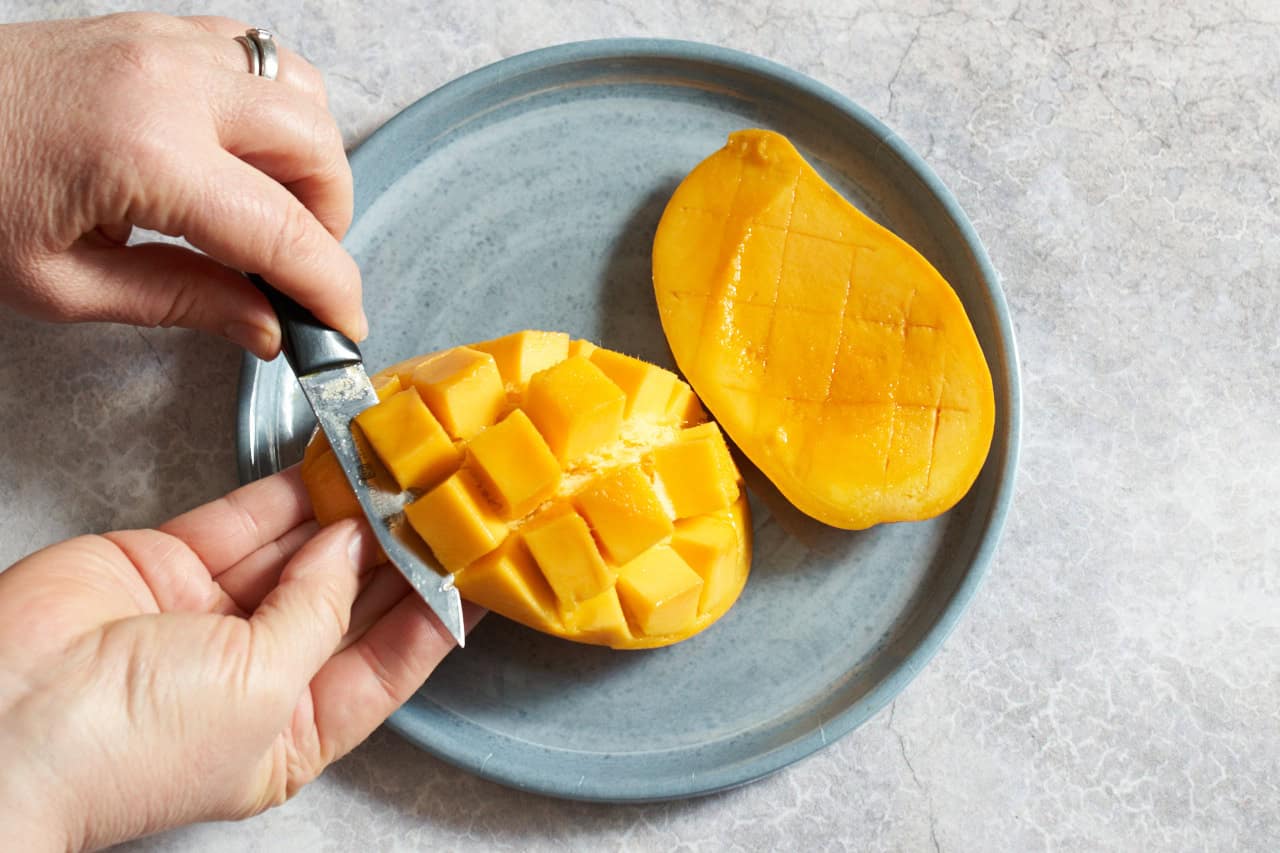 A woman's hands shown cutting a mango that has been cross-hatched into chunks away from the peel, another mango half is on the right.